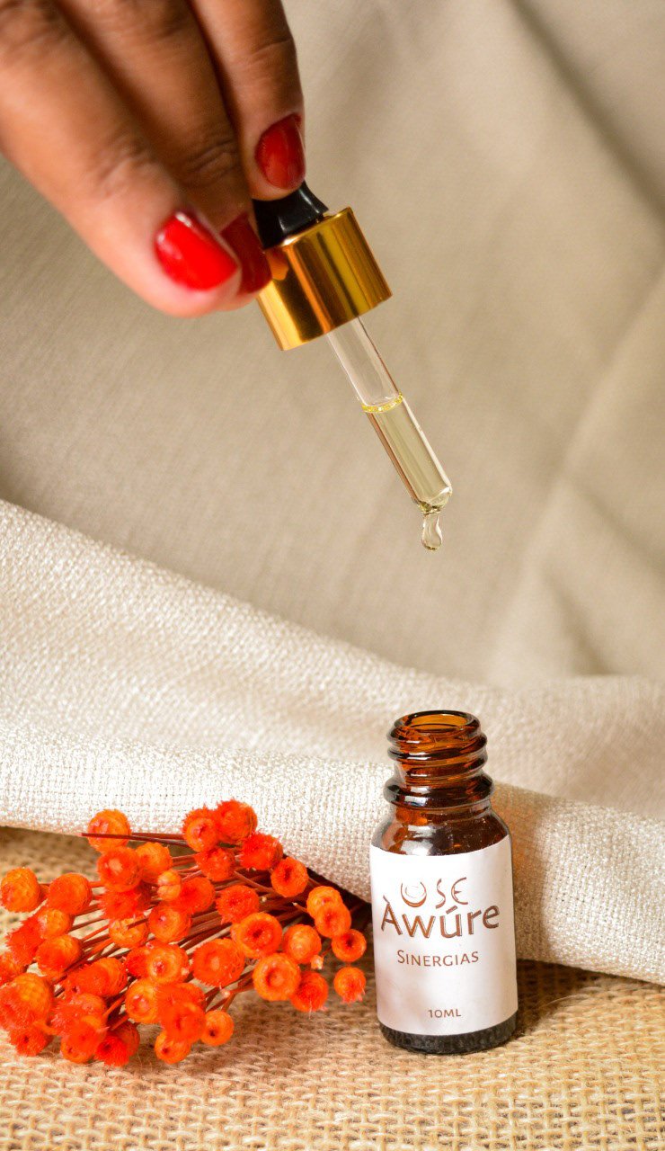 Hand with red nails holding a golden dropper over the USE Àwúre Sinergias amber bottle with dried orange flowers